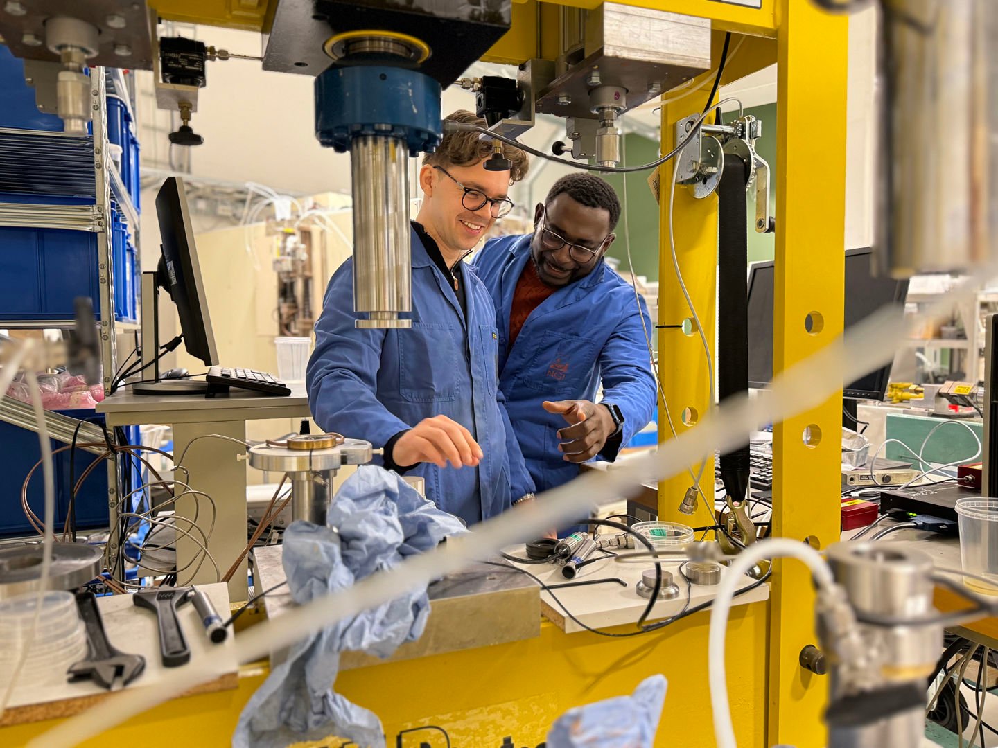 Fredrik og Jacob, iført hver sin blå laboratoriefrakk, står i et mekanisk laboratorium og kikker på en gul maskin.